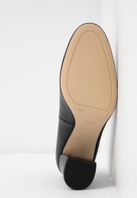 Chaussure en cuir noir avec semelle beige, talon bloc bas, taille 4½, et marque "högl" sur la semelle, placée à la verticale contre un mur blanc d’angle.
