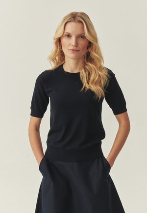 Lyshåret kvinde med bølget hår iført sort kortærmet top og nederdel, stående med hænderne i lommerne mod ensfarvet baggrund.