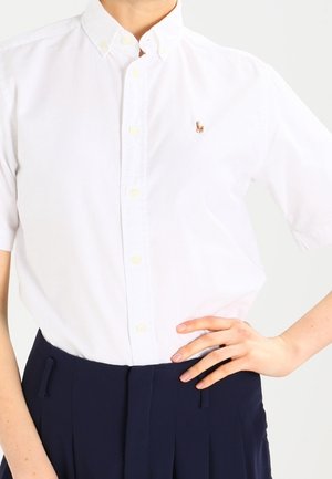 Osoba ubrana w biały krótki rękaw koszuli na guziki z małym wyszywanym logo oraz ciemne, wysokie spodnie, z ręką na biodrze.