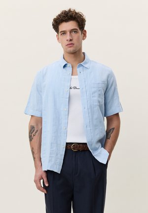 Ung mand med krøllet hår iført lyseblå kortærmet skjorte over hvidt tanktop og mørke bukser, stående med den ene hånd i lommen.