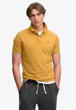 Jeune homme portant un polo jaune moutarde à manches courtes par-dessus un maillot blanc, avec un pantalon gris foncé à cordon, mains dans les poches.