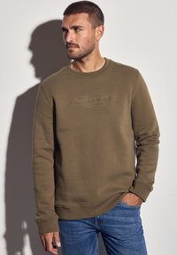 Mand med kort hår og skæg iført olivengrøn sweatshirt og blå jeans, stående med den ene hånd i lommen, kiggende til venstre.