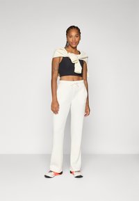 Een vrouw staat zelfverzekerd, gekleed in een zwarte cropped top, witte broek en sportschoenen, met een lichte sweater over haar schouders.