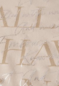 Tissu beige avec des motifs de texte en argent en relief. Présente à la fois des lettres en gras et en cursive, ajoutant texture et intérêt visuel.