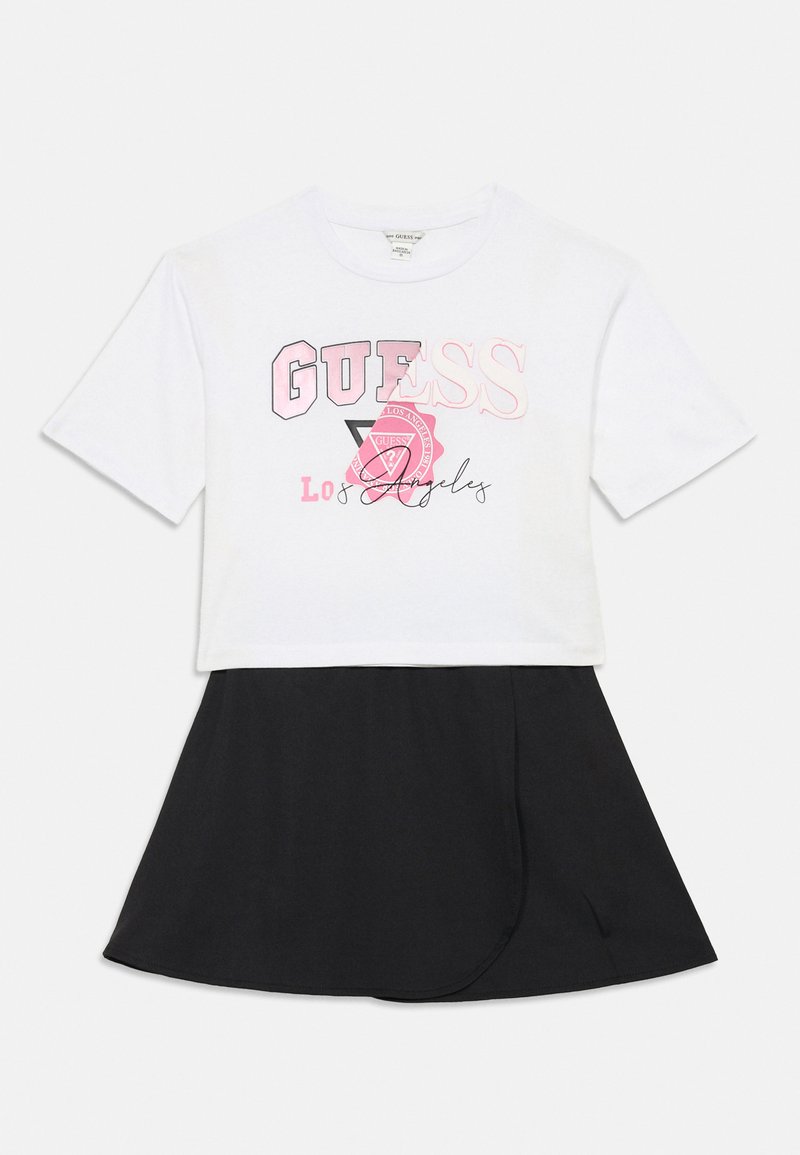 Maglietta bianca corta con grafica "GUESS Los Angeles" in rosa e nero, abbinata a una gonna nera con un design drappeggiato.