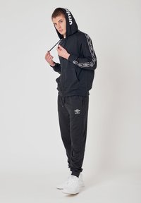 Felpa nera con zip e logo bianco, accentuata da dettagli a fantasia, abbinata a pantaloni jogger neri con logo. Indossata con sneakers bianche.