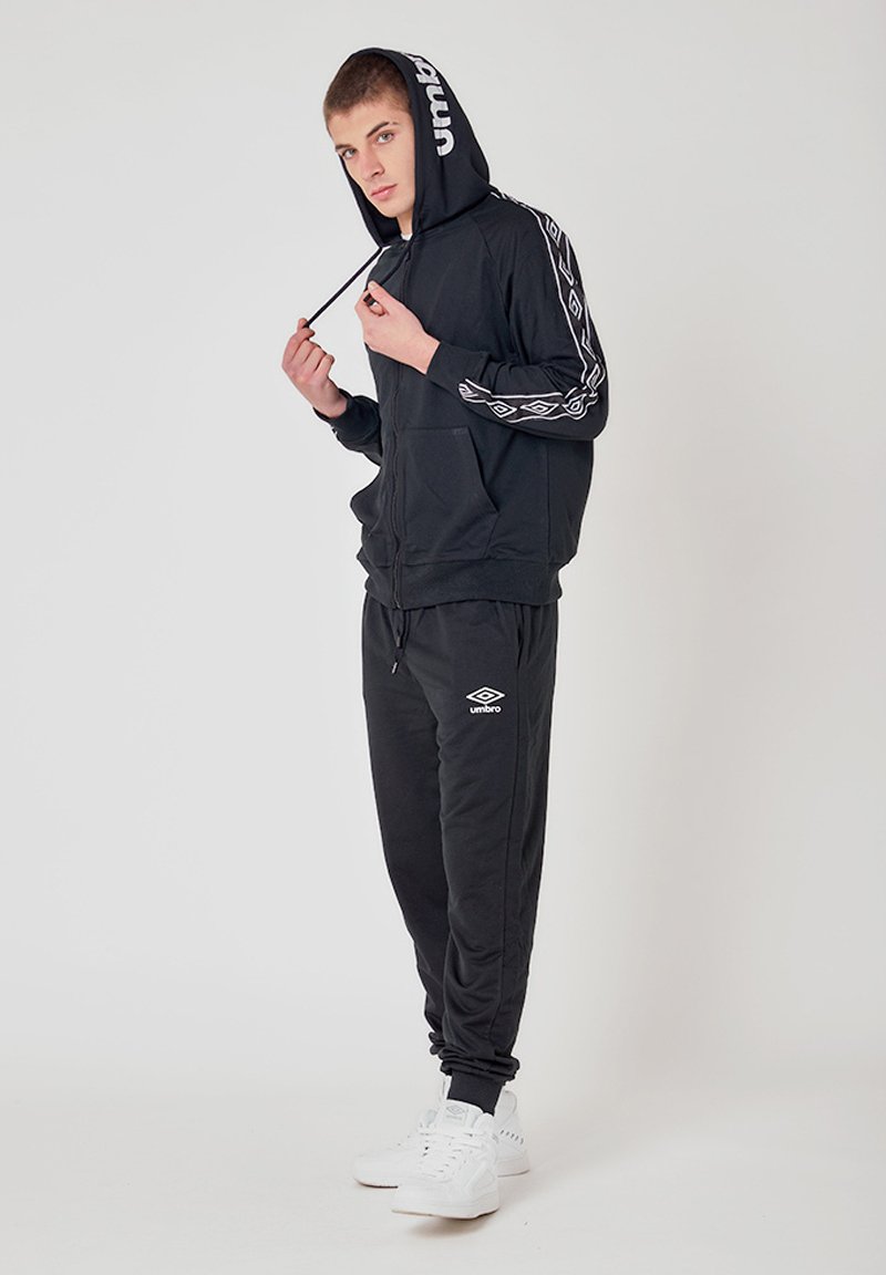 Felpa nera con zip e logo bianco, accentuata da dettagli a fantasia, abbinata a pantaloni jogger neri con logo. Indossata con sneakers bianche.