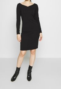 Svart långärmad klänning med V-ringning, figurnära siluett och knälång längd, kombinerad med svarta texturerade ankelskor.