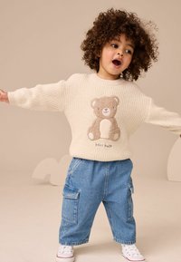 Maglione lavorato a maglia color crema con un'illustrazione di un orsetto, abbinato a pantaloni cargo blu. Le sneaker del bambino sono bianche con accenti rossi.