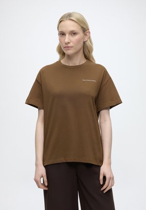 Jeune femme aux cheveux blonds porte un t-shirt marron Marc O’Polo Denim et un pantalon foncé, debout devant un fond clair uni.