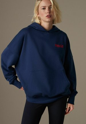 Granatowy hoodie wykonany z miękkiego materiału, z kieszenią kangurzą i czerwonym logo "Coca-Cola" na klatce piersiowej. Luźny krój i kaptur ze sznurkiem.