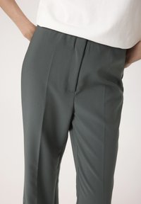 Krojene hlače temno zelene barve iz gladkega materiala z visokim pasom, ki imajo en sam sprednji plis in stranske žepe.