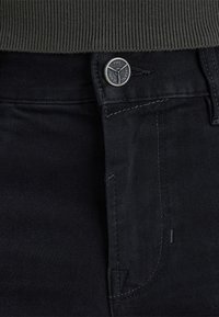 Primo piano della vita con la cintura dei jeans in denim nero, con un evidente bottone di metallo inciso "PME Legend Jeans" e la parte superiore di un maglione verde scuro a coste infilato.