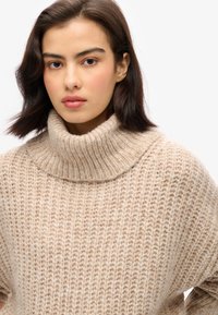 Suéter de punto beige con un cuello alto y acanalado. La textura es suave, con un corte holgado y sutiles patrones horizontales en todo el tejido.