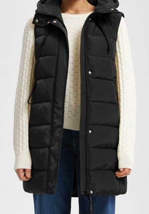 Woman wearing a sleeveless black quilted vest over a white textured long-sleeve sweater and blue jeans, standing with arms relaxed.