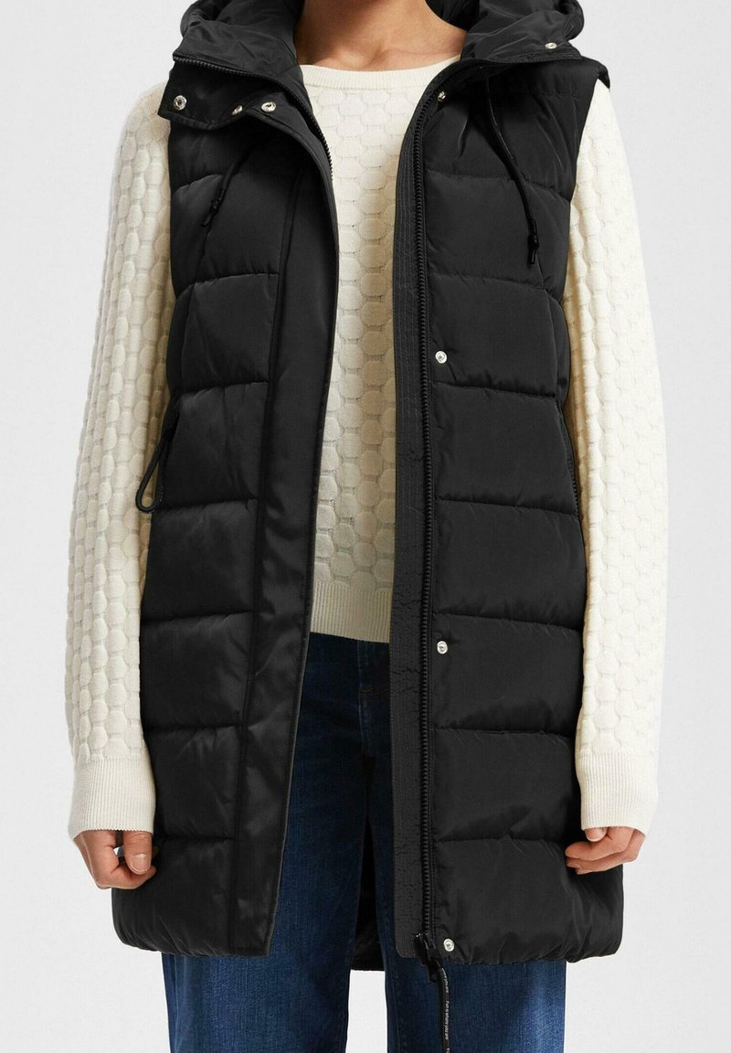 Femme portant un gilet matelassé noir sans manches par-dessus un pull blanc texturé à manches longues et un jean bleu, debout, les bras détendus.