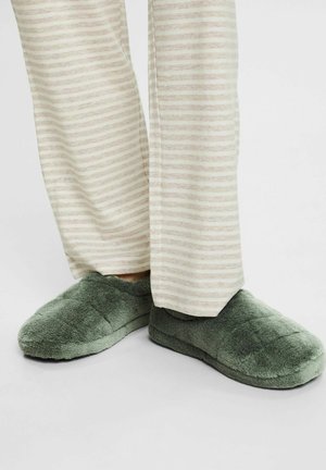 Fødder iført bløde, grønne plyssede hjemmesko og beige stribede pyjamasbukser står på en almindelig hvid overflade.
