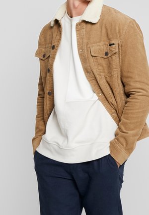 Homme portant une veste en velours côtelé beige avec col en polaire blanc, sweat-shirt blanc et pantalon bleu foncé, mains dans les poches, sur fond uni.