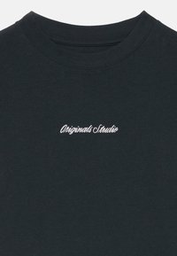 T-shirt en coton noir avec un col rond et une broderie cursive blanche portant l'inscription "Originals Studio" sur la poitrine. Texture douce, design décontracté.