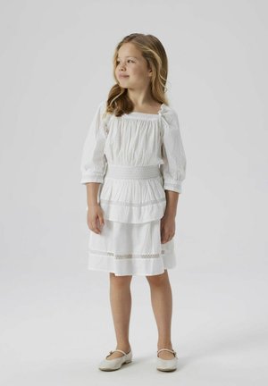 Witte jurk met opzettige mouwen, smocktail, gelaagde rok en subtiele textuurdetails. Gecombineerd met beige schoenen.