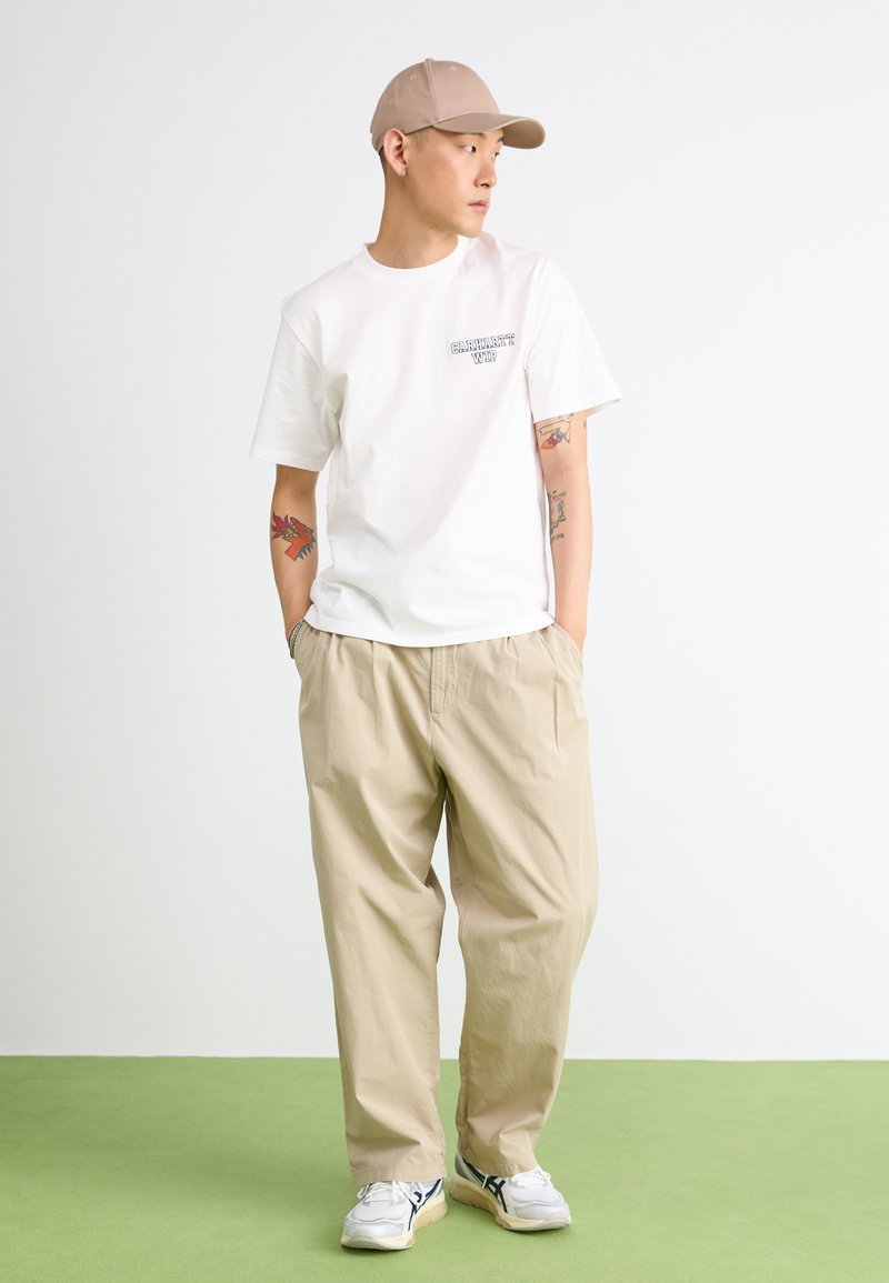 T-shirt blanche à manches courtes avec un graphique, pantalon ample beige, casquette beige et baskets blanches avec des accents bleus sur un sol vert.