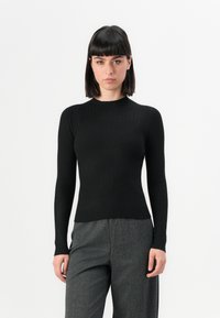 Donna con capelli scuri e lisci, che indossa un top a maniche lunghe nero a costine aderente e pantaloni grigi con texture, in piedi davanti a uno sfondo bianco.