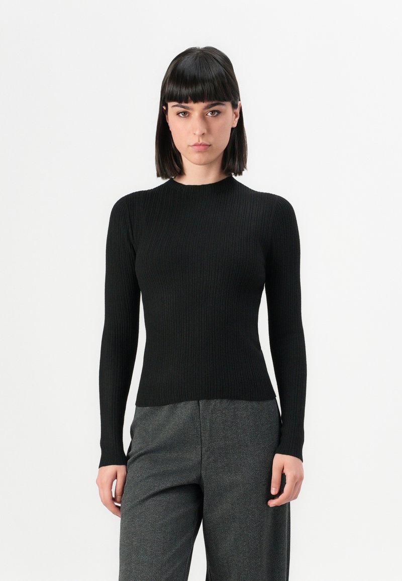 Donna con capelli scuri e lisci, che indossa un top a maniche lunghe nero a costine aderente e pantaloni grigi con texture, in piedi davanti a uno sfondo bianco.