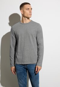 Graues Langarm-T-Shirt aus weichem Material mit Rundhalsausschnitt, lockerer Passform und dezentem Logo auf der Brust. Getragen mit blauen Jeans.