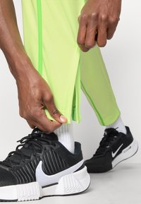 Pantalones deportivos verde lima con cremalleras laterales, combinados con zapatillas negras que tienen un logo blanco y acentos texturizados, y calcetines blancos.