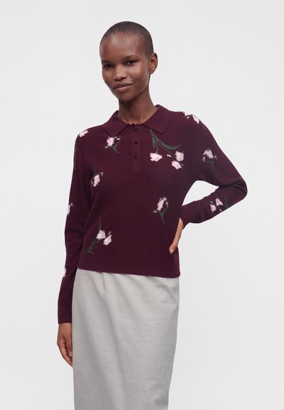 Personne portant un pull à col violet foncé avec un motif floral rose et une jupe gris clair, debout avec une main sur la hanche devant un fond uni.
