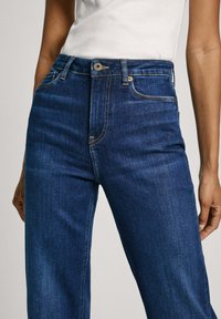 Modré džíny s vysokým pasem vyrobené z denimu, které mají klasický design s pěti kapsami, kontrastní prošívání a jediné zapínání na knoflík v pase.