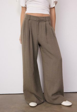 Braune, hoch taillierte, weite Hosen mit Falten, präsentiert mit einem weißen, kurz geschnittenen Oberteil und weißen Schuhen, stehend vor einer schlichten Wand.