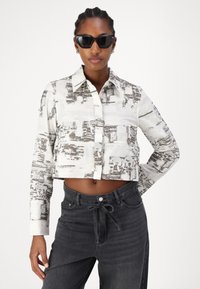 Kuttet skjorte i hvit silke med et svart abstrakt mønster, med krage og lange ermer, kombinert med mørk grå jeans med snøring.
