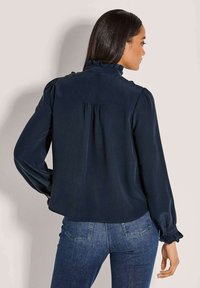 Dunkelbluse mit langen Ärmeln, Rüschensaum und akzentuierten Schultern. Die Rückseite hat eine Falten-Detail. Glatte Stofftextur.