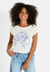 Camiseta blanca de manga corta con un diseño de cara dibujada en líneas azules, con cuello redondo y corte relajado, combinada con pantalones de color azul claro.