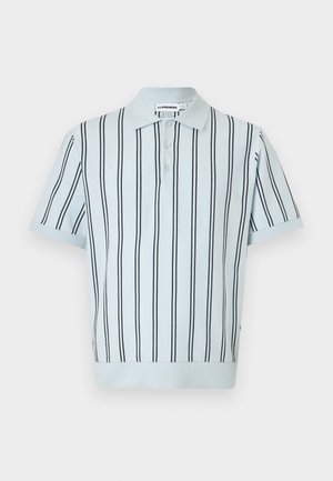 Mann trägt ein hellblaues, kurzärmeliges, gestreiftes Hemd, marineblaue Shorts, schwarze Sandalen, hält eine schwarze Tasche unter dem Arm und steht vor schlichtem Hintergrund.