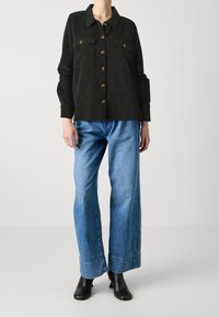 Černá kordurová košile na knoflíky se dvěma předními kapsami, kombinovaná s volnými modrými džíny a černými kotníkovými botami.