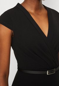 Femme portant une robe noire style portefeuille à manches courtes et une fine ceinture noire avec une boucle argentée à la taille.