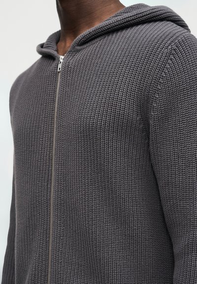 Άνδρας που φοράει σκούρο γκρι, πλεκτό φούτερ με φερμουάρ, με το φερμουάρ εντελώς κλειστό, εμφανιζόμενος από τον λαιμό μέχρι το μέσο του κορμού.