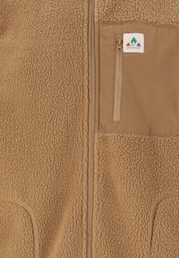 Gilet en polaire beige avec une surface texturée, comportant une poche poitrine zippée et des détails en tissu lisse. Patch logo sur la poche.