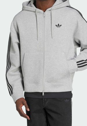 Mand iført lysegrå Adidas lynlås-hættetrøje med sorte tre striber på ærmerne og sort Adidas-logo, kombineret med sorte bukser.