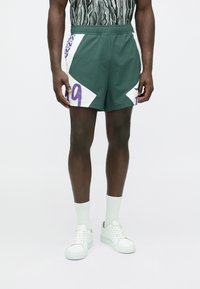 Zöld sportnadrág fehér és lila díszítéssel; az oldalakon lila "19" szám és geometriai mintázat található.