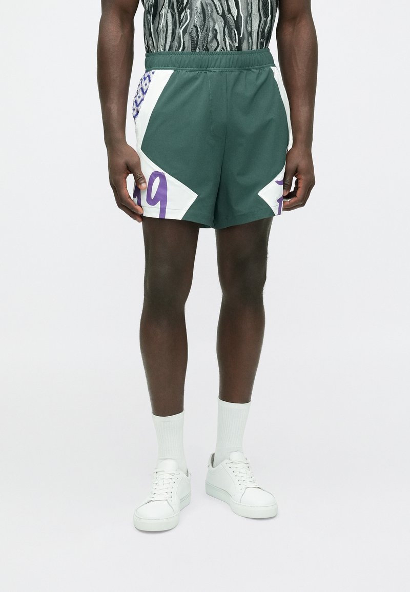 Zöld sportnadrág fehér és lila díszítéssel; az oldalakon lila "19" szám és geometriai mintázat található.