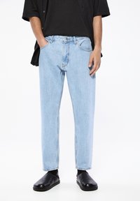 Blugi din denim de culoare albastru deschis cu o croială lejeră, cinci buzunare și detalii de cusătură. Purtați cu un tricou negru și pantofi slip-on negri.