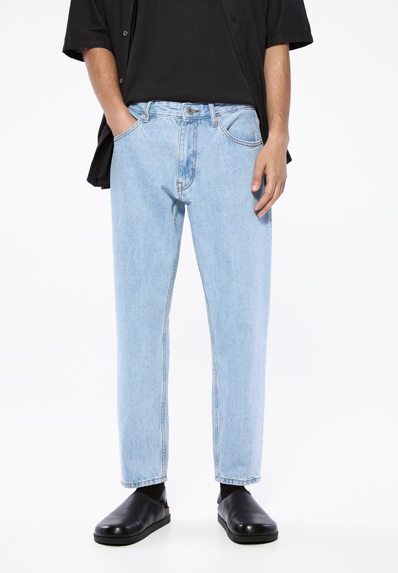 Blugi din denim de culoare albastru deschis cu o croială lejeră, cinci buzunare și detalii de cusătură. Purtați cu un tricou negru și pantofi slip-on negri.