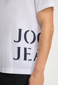 Weißes Baumwoll-T-Shirt mit fettem schwarzen Aufdruck "JOG JEAN" auf der linken Seite. Kurze Ärmel und eine lockere Passform mit glatter Textur.