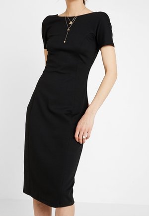 Kobieta ubrana w dopasowaną czarną sukienkę za kolano z krótkimi rękawami, ozdobioną warstwami złotych naszyjników i pierścionkiem na lewej dłoni.