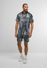 Sort og grå mønstret sportsoutfit bestående af en kortærmet skjorte og shorts, med et logo på brystet og et mærke på benet. Hvide sneakers.