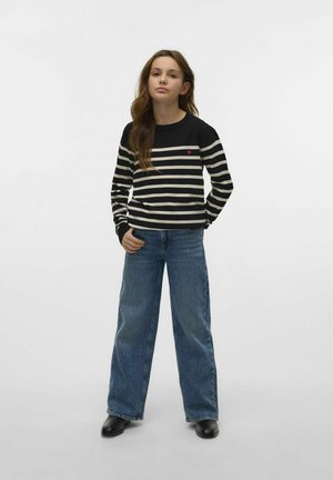Jeune femme debout, les mains dans les poches, portant un pull rayé noir et blanc avec un cœur rouge et un jeans bleu à pattes larges.