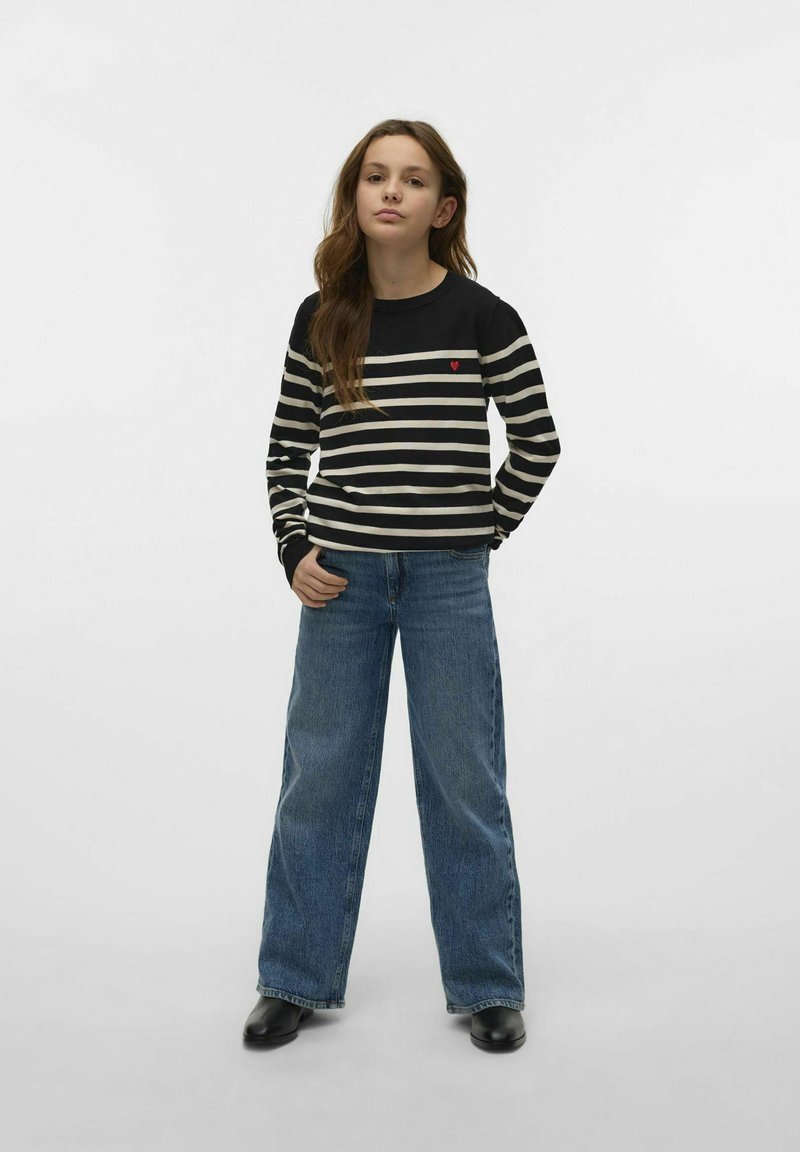 Mlada ženska stoji z rokami v žepih, oblečena v črno-belo črtasto pulover s rdečim srcem in široke modre jeans hlače.